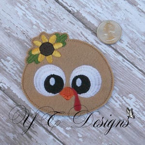 May include: A brown felt appliqu&eacute; of a turkey with a yellow sunflower on its head. The turkey has large white eyes with black pupils and a red wattle.