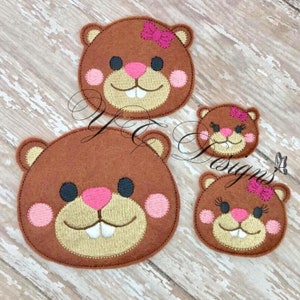 May include: Three felt appliques of brown bear faces with pink cheeks and noses. The largest bear has a pink bow on its head. The smallest bear has eyelashes.
