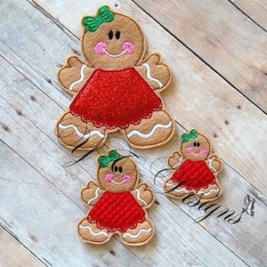 May include: Three felt gingerbread people ornaments. The largest gingerbread person is wearing a red dress with white trim and a green bow. The two smaller gingerbread people are wearing red dresses with white trim and green bows. The ornaments are on a wooden background.