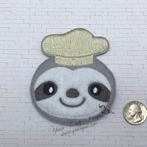 Puede incluir: Un perezoso de fieltro gris que lleva un gorro de chef de fieltro amarillo. El perezoso tiene ojos, nariz y boca bordados.