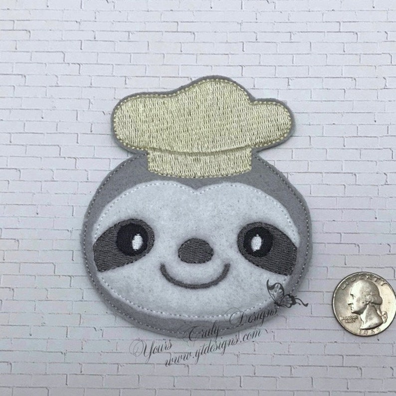 Puede incluir: Un perezoso de fieltro gris que lleva un gorro de chef de fieltro amarillo. El perezoso tiene ojos, nariz y boca bordados.