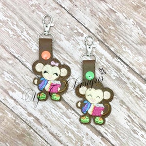 Może przedstawiać: Dwa brązowe breloczki w kształcie małpy z kolorowymi plecakami i książkami. Małpy trzymają książki i noszą kolorowe buty. Breloczki mają srebrny klips i brązowy pasek ze skóry.