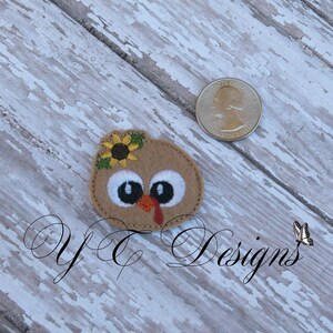 May include: A brown felt applique of a turkey with a sunflower on its head. The turkey has large white eyes and a red wattle.