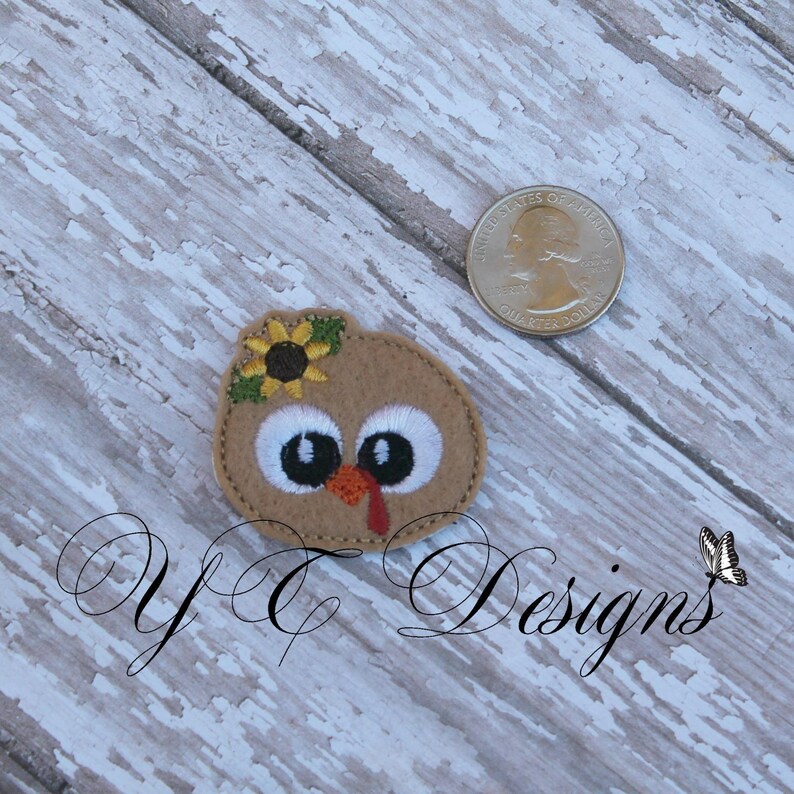 May include: A brown felt applique of a turkey with a sunflower on its head. The turkey has large white eyes and a red wattle.
