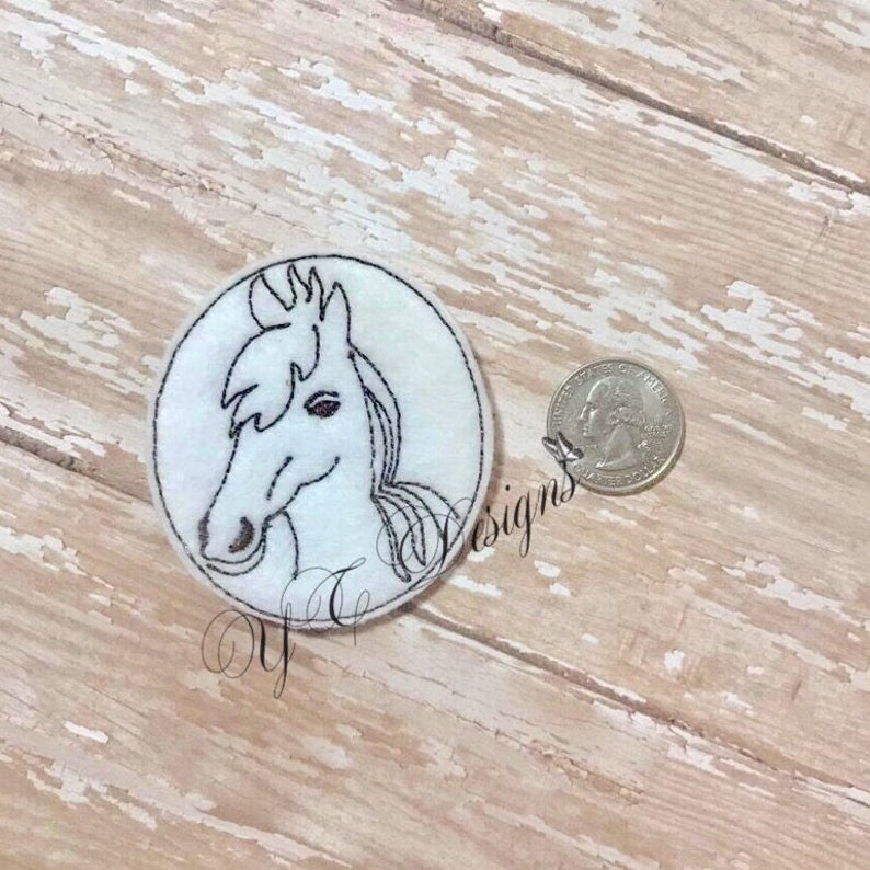 Puede incluir: Una aplicaci&oacute;n de fieltro blanco de la cabeza de un caballo con detalles bordados en negro. La aplicaci&oacute;n es circular y del tama&ntilde;o aproximado de una moneda de 25 centavos de d&oacute;lar estadounidense.