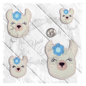 Puede incluir: Cuatro caras de llama bordadas con los ojos cerrados y una flor azul en la cabeza. Las llamas son blancas con narices rosas y están sobre un fondo de madera blanco.