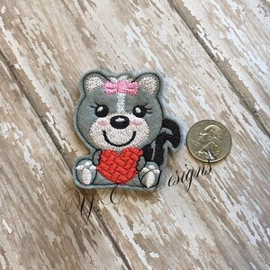 Puede incluir: Una aplicaci&oacute;n bordada gris de un animal de dibujos animados que sostiene un coraz&oacute;n rojo. El animal tiene un lazo rosa en la cabeza y ojos negros.