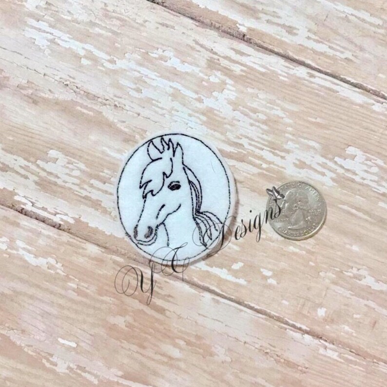 Puede incluir: Un c&iacute;rculo de fieltro blanco con un dibujo de l&iacute;nea negra de la cabeza de un caballo. El caballo tiene una crin y una cola.