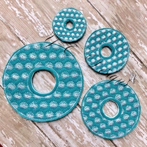 Puede incluir: Tres círculos de fieltro azul turquesa con diseños bordados en blanco. Los círculos tienen un agujero en el centro y son de diferentes tamaños.