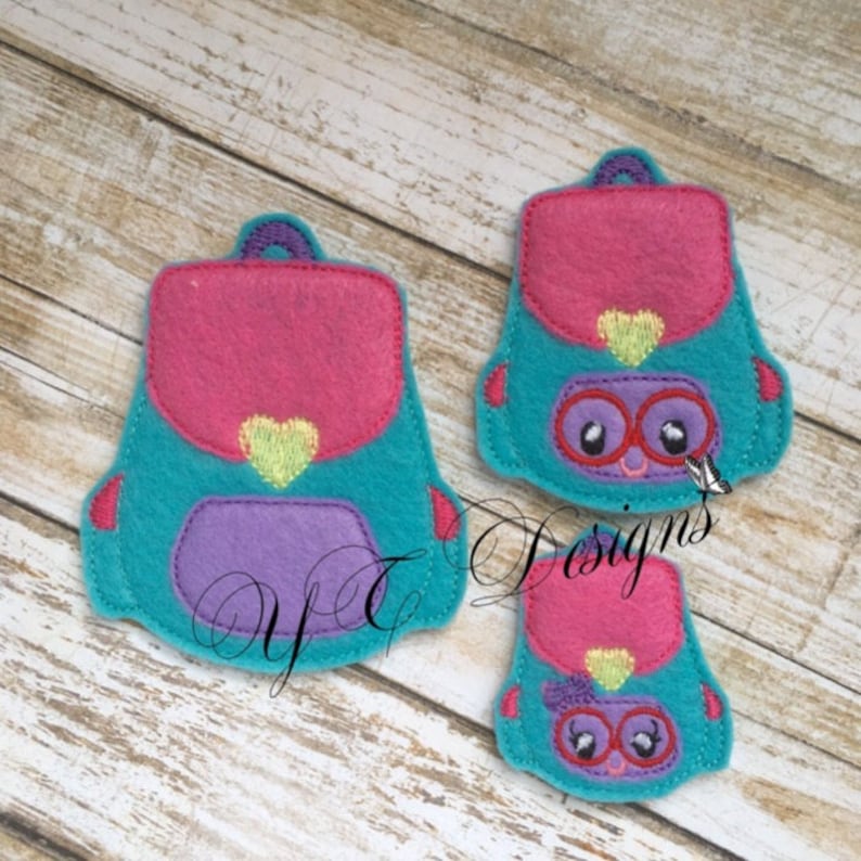May include: Two felt backpack appliques with a pink, purple, and teal color scheme. The larger applique has a yellow heart and the smaller applique has a yellow bow. Both appliques have a pair of glasses and a smiling face.