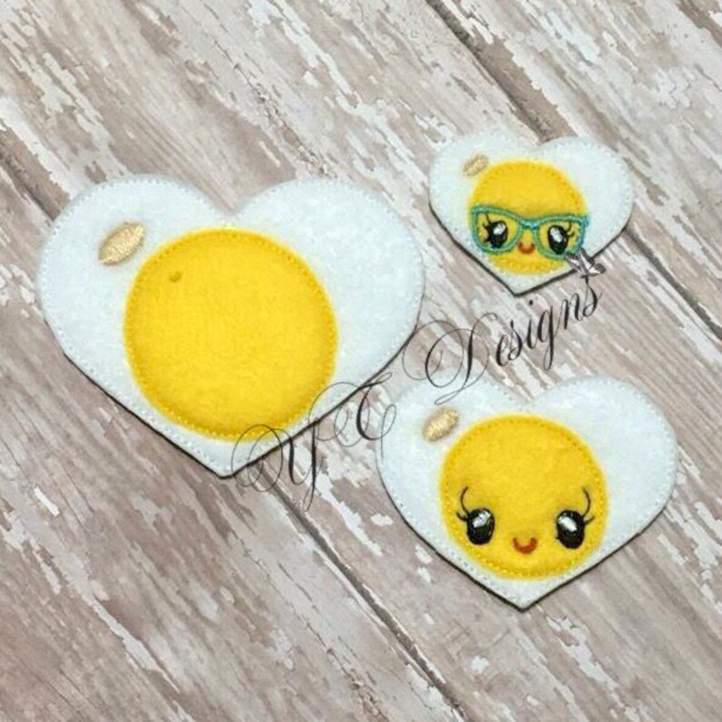 May include: Three felt heart-shaped patches with a fried egg design. The eggs have yellow yolks and white whites. One egg has a smiling face with eyelashes, another has a smiling face with sunglasses, and the third egg has no face.