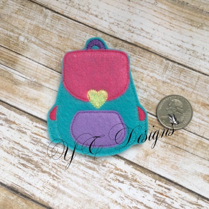 May include: A small felt backpack with a pink top flap, a purple pocket, and a green heart on the front. The backpack is about the size of a US quarter.