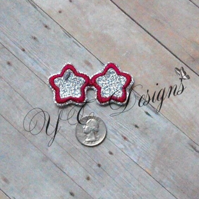 May include: Star-shaped sunglasses with a red border and silver glitter filling. The sunglasses are placed on a wooden surface next to a quarter. The text "yC designs" is visible in the image.