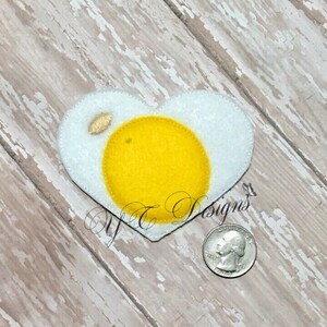 May include: A felt heart-shaped patch in the shape of a fried egg. The yolk is yellow and the white is white.