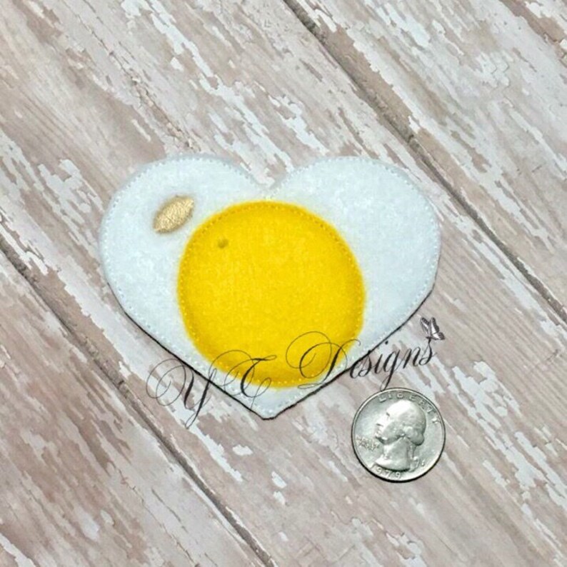 May include: A felt heart-shaped patch in the shape of a fried egg. The yolk is yellow and the white is white.