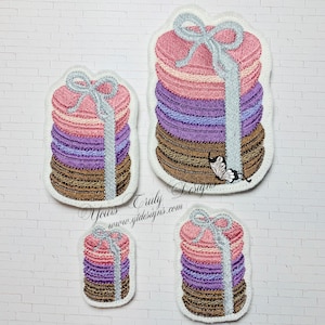 May include: Three embroidered patches of stacked macarons tied with a ribbon. The largest patch is 8.9 cm tall, the medium patch is 6.4 cm tall, and the smallest patch is 3.8 cm tall. The patches are in shades of pink, purple, and brown.