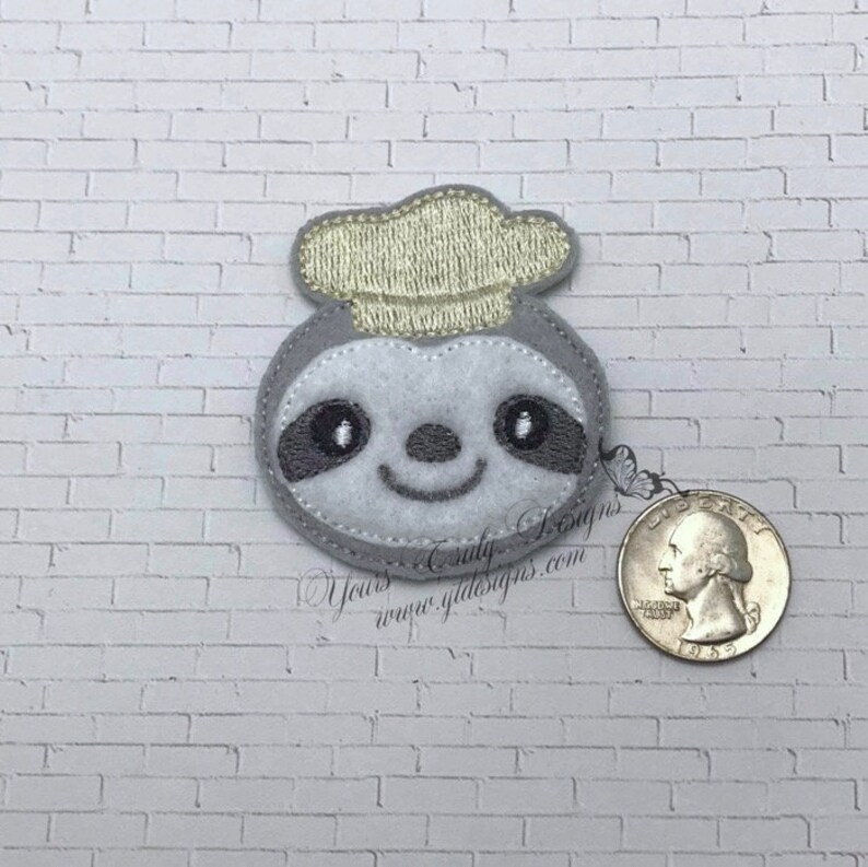 Puede incluir: Un perezoso de fieltro gris que lleva un gorro de chef amarillo. El perezoso tiene una cara sonriente con ojos negros y una nariz gris. El perezoso es aproximadamente del tama&ntilde;o de una moneda de 25 centavos.