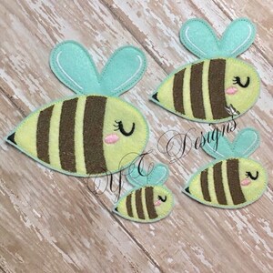 May include: Three felt bee appliques in different sizes. The bees are yellow with brown stripes and have light blue wings. The bees have black eyes and pink cheeks.