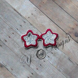 May include: Star-shaped sunglasses with a red border and silver glitter filling. The glasses are on a wooden surface. The glasses are a fun accessory.