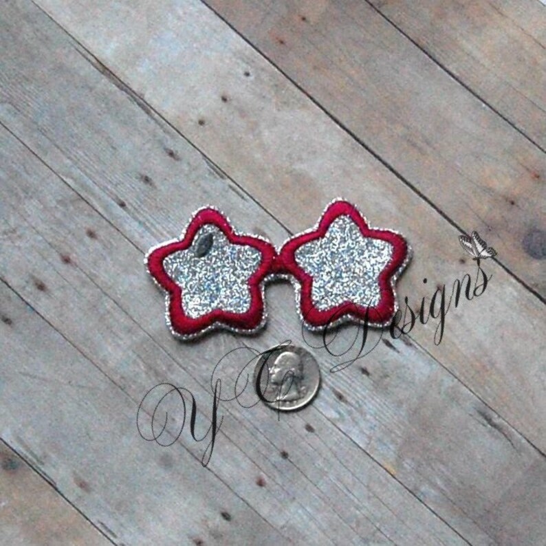 May include: Star-shaped sunglasses with a red border and silver glitter filling. The glasses are on a wooden surface. The glasses are a fun accessory.