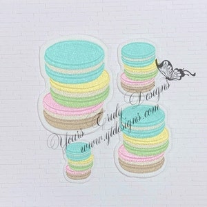 May include: Three embroidered patches of pastel-coloured macarons. The largest macaron is stacked with five layers of light blue, yellow, green, pink, and brown. The middle macaron is stacked with four layers of light blue, yellow, green, and pink. The smallest macaron is stacked with four layers of light blue, yellow, green, and pink.