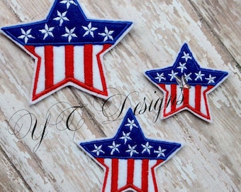 Pode incluir: Três patches bordados em forma de estrela em vermelho, branco e azul, que se assemelham à bandeira americana. As estrelas são de tamanhos diferentes, a maior na parte superior e a menor na parte inferior.
