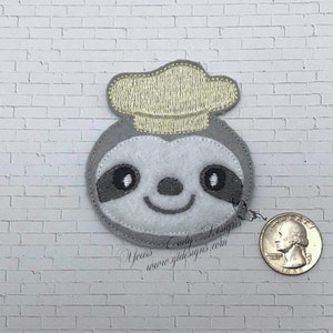 Puede incluir: Un perezoso de fieltro gris que lleva un gorro de chef de fieltro amarillo. El perezoso tiene ojos negros y una sonrisa cosida. El perezoso tiene aproximadamente 5 cm de di&aacute;metro.