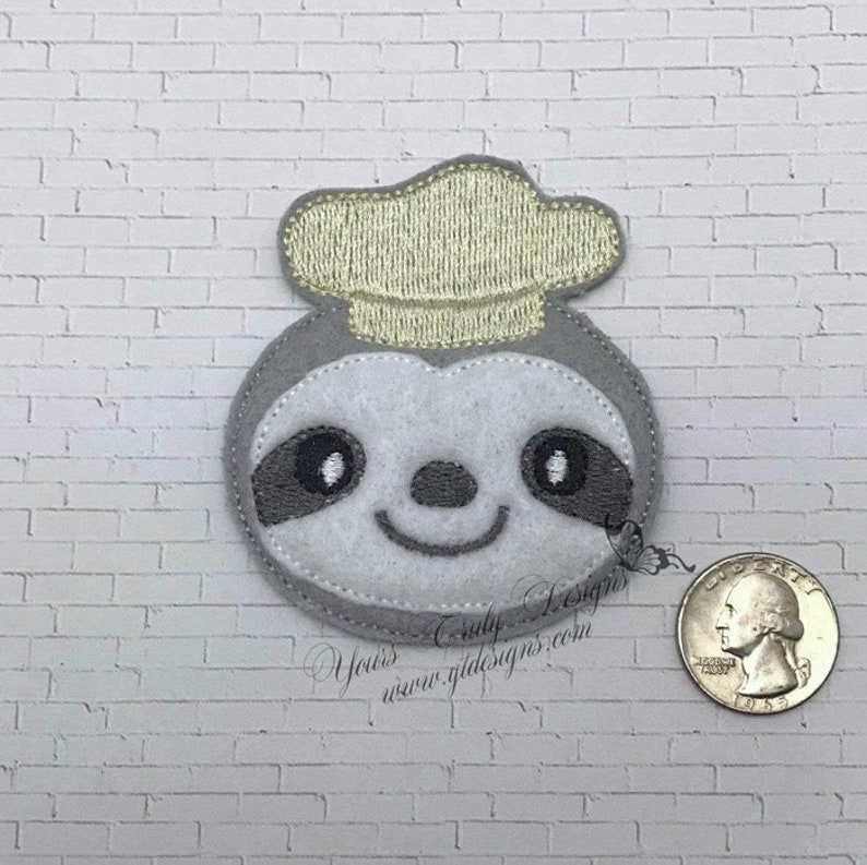 Puede incluir: Un perezoso de fieltro gris que lleva un gorro de chef de fieltro amarillo. El perezoso tiene ojos negros y una sonrisa cosida. El perezoso tiene aproximadamente 5 cm de di&aacute;metro.