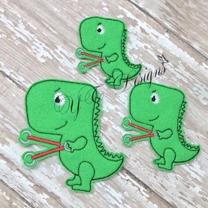 May include: Three green felt dinosaur appliques with red and white embroidery. Each dinosaur is holding a pair of red and white dumbbells.