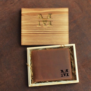 Może przedstawiać: Brązowy sk&oacute;rzany portfel z personalizowanym monogramem w drewnianym pudełku. Monogram to litera "M" z imieniem "Max" poniżej. Pudełko ma drewnianą pokrywę z tym samym monogramem i tekstem "Wszystkiego najlepszego z okazji urodzin!"