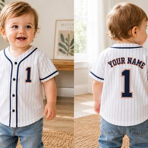 Pode incluir: Uma camisola de basebol branca com riscas verticais finas, acabamentos em azul escuro e fecho de botões. A frente apresenta o número 1 em azul escuro com contornos laranja. As costas exibem o texto YOUR NAME e o número 1 em azul.