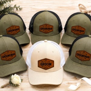 Può includere: Cinque cappellini da baseball disposti in gruppo. Quattro cappellini sono verde oliva con retro in rete nera e toppe in similpelle marrone con la scritta GROOMSMAN. Un cappellino centrale è color crema con rete bianca e toppa GROOM.