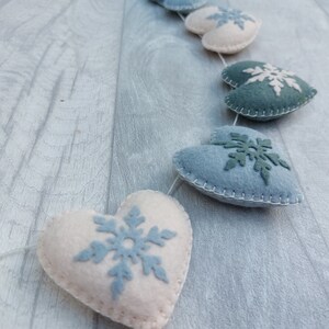 May include: A garland of felt hearts in white, blue, and teal. Each heart features a snowflake design in a contrasting colour. The hearts are strung together with a thin white thread, creating a festive winter decoration.