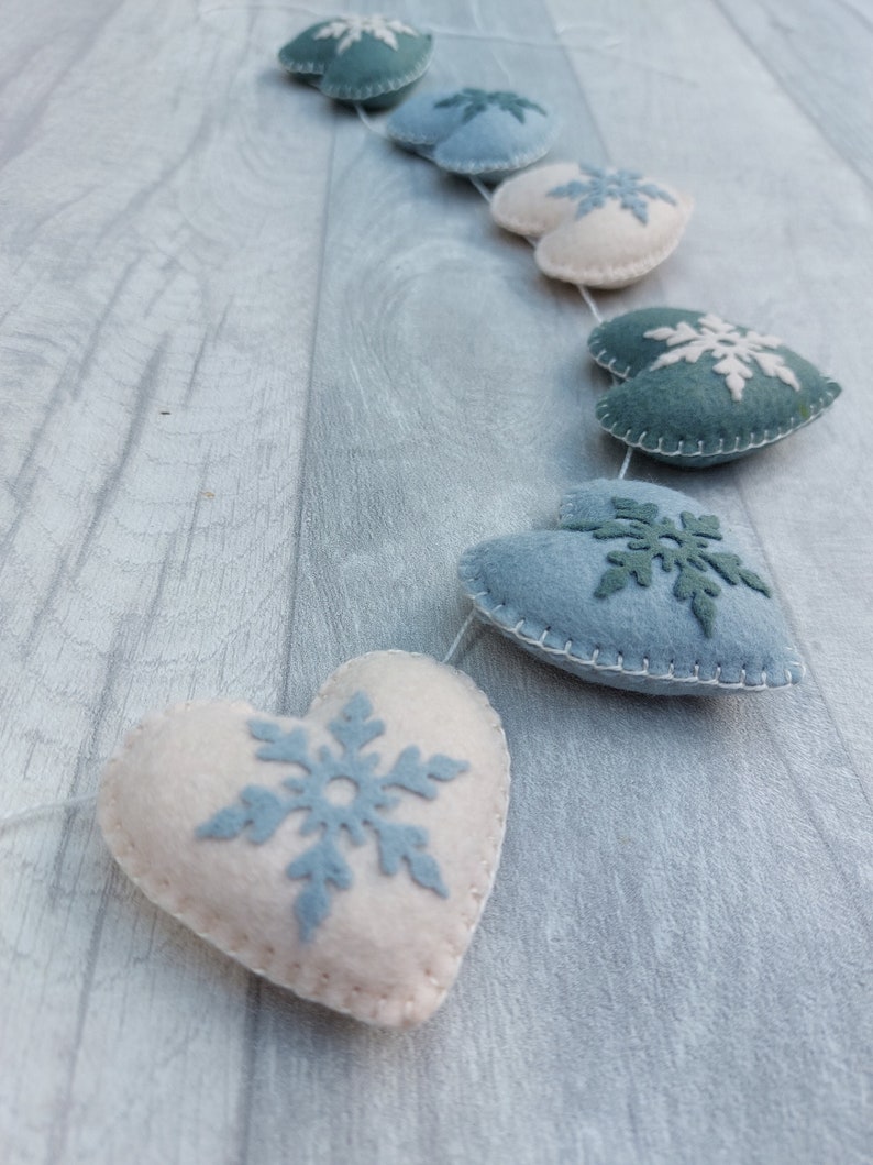 May include: A garland of felt hearts in white, blue, and teal. Each heart features a snowflake design in a contrasting colour. The hearts are strung together with a thin white thread, creating a festive winter decoration.