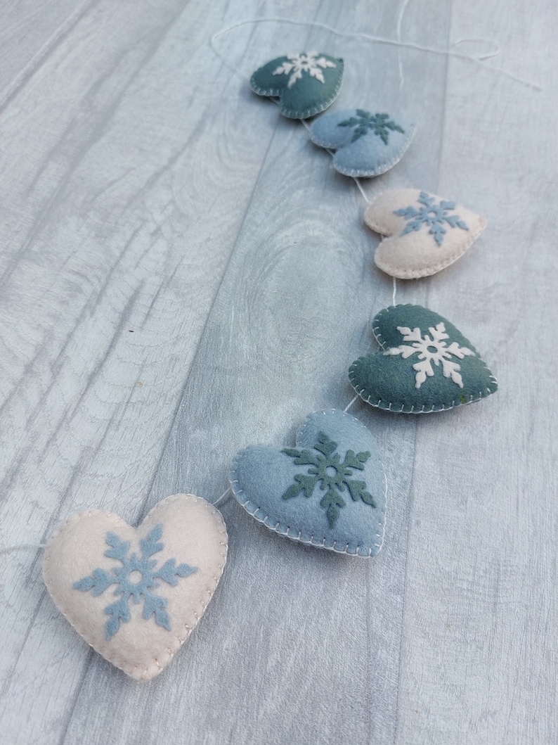 May include: A garland of felt hearts in various shades of blue and cream, each featuring a snowflake design. The hearts are connected by white thread, creating a decorative item. The garland is ideal for festive occasions.