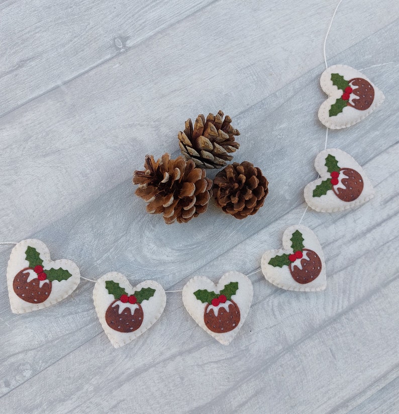May include: A white felt garland with green holly leaves and red berries. Each heart-shaped ornament features a brown felt Christmas pudding with white icing.