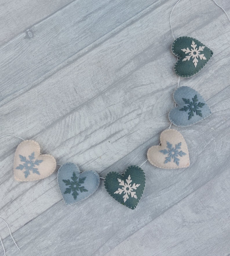 May include: A garland of felt hearts in cream, blue, and green, each decorated with a snowflake. The hearts are strung together with white thread, creating a festive winter decoration for the home.