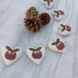 May include: A white felt garland with red and green felt Christmas pudding ornaments in the shape of hearts. The garland is strung with white twine and is hanging on a white wooden surface.
