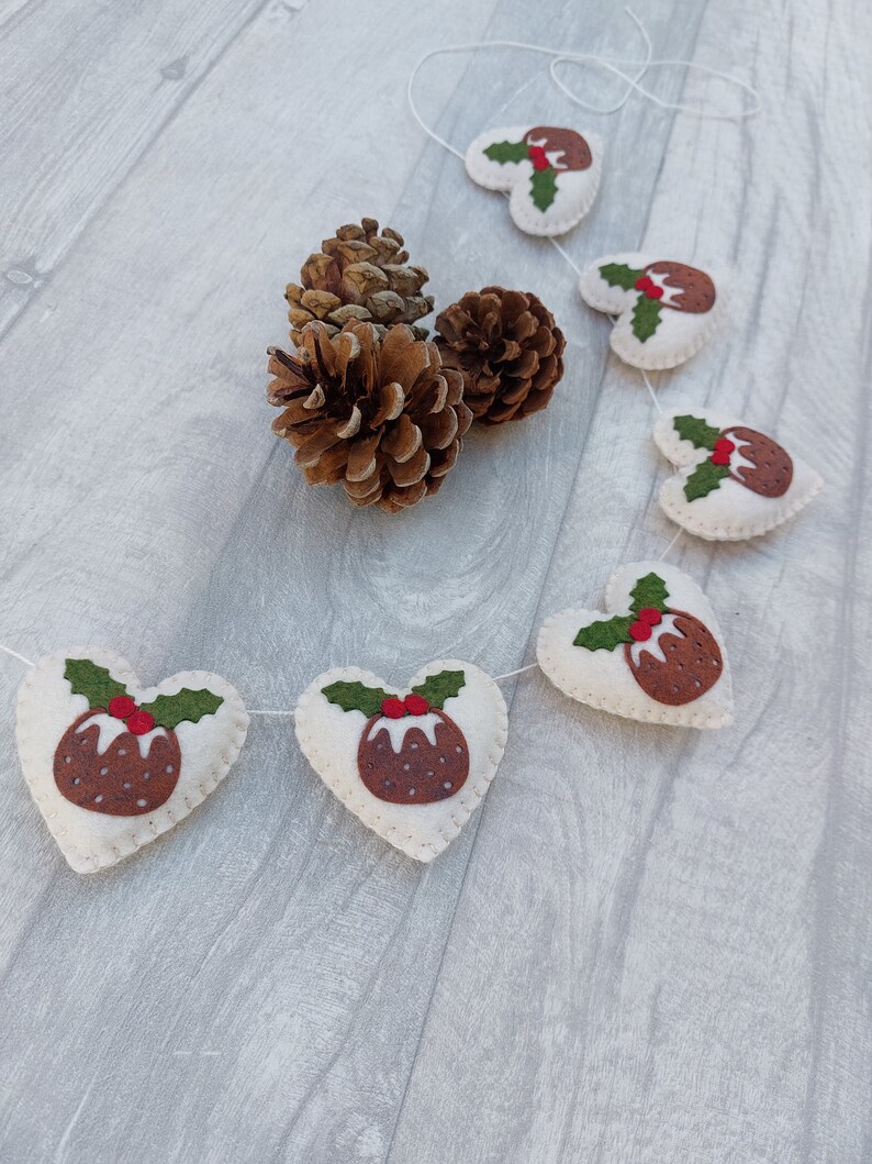 May include: A white felt garland with red and green felt Christmas pudding ornaments in the shape of hearts. The garland is strung with white twine and is hanging on a white wooden surface.