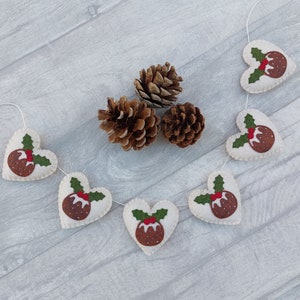 May include: A white felt garland with red and green felt Christmas pudding ornaments in the shape of hearts. The garland is strung with white thread and is decorated with pine cones.