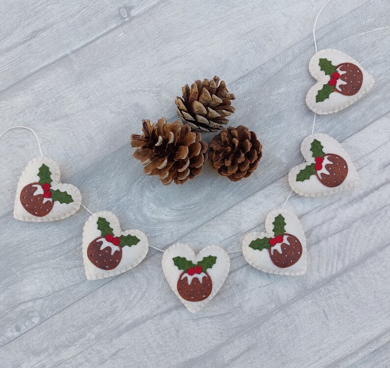 May include: A white felt garland with red and green felt Christmas pudding ornaments in the shape of hearts. The garland is strung with white thread and is decorated with pine cones.