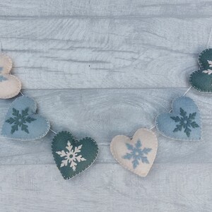 May include: A garland of felt hearts in shades of blue, green, and cream, each decorated with a snowflake design. The hearts are strung together with white thread, creating a festive winter decoration.