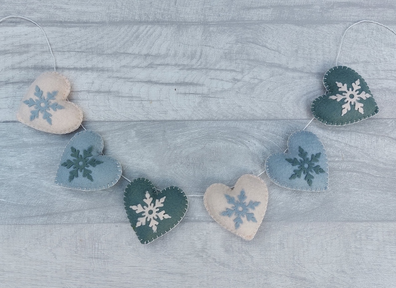 May include: A garland of felt hearts in shades of blue, green, and cream, each decorated with a snowflake design. The hearts are strung together with white thread, creating a festive winter decoration.