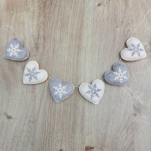 May include: A garland of six white felt hearts with grey snowflake designs, strung together with white thread. The garland is hanging against a light brown wood background.