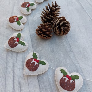 May include: A white felt garland with brown and green felt Christmas pudding ornaments. The garland is strung with white thread and is hanging on a light wood background.