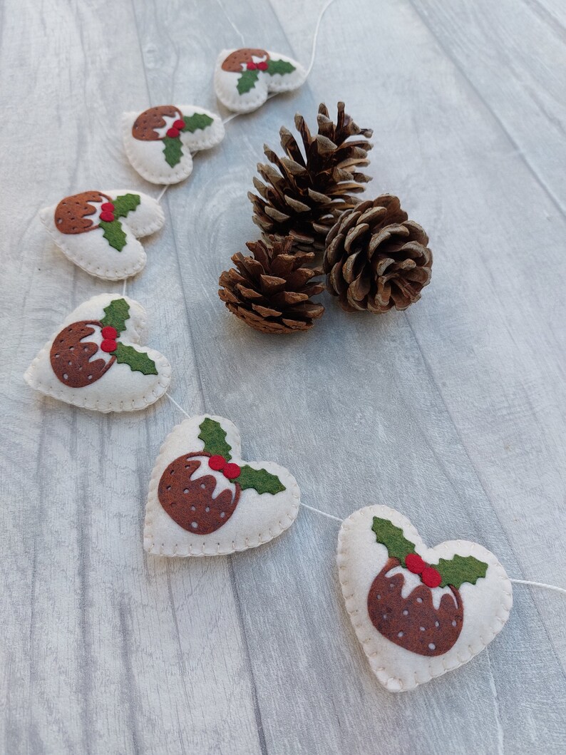 May include: A white felt garland with brown and green felt Christmas pudding ornaments. The garland is strung with white thread and is hanging on a light wood background.