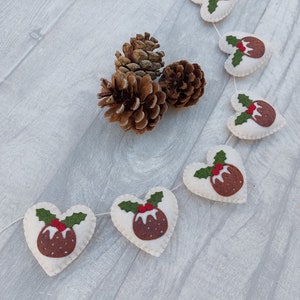 May include: A white felt Christmas garland with red and green felt Christmas pudding ornaments in the shape of hearts. The garland is strung with white thread and is hanging on a white wooden surface.