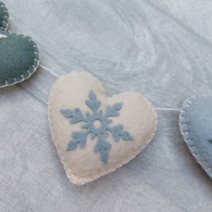 May include: A festive garland featuring felt hearts in cream and teal, stitched with white thread. Each heart is adorned with a light blue snowflake. The garland is strung on a white string, ideal for winter decorations.