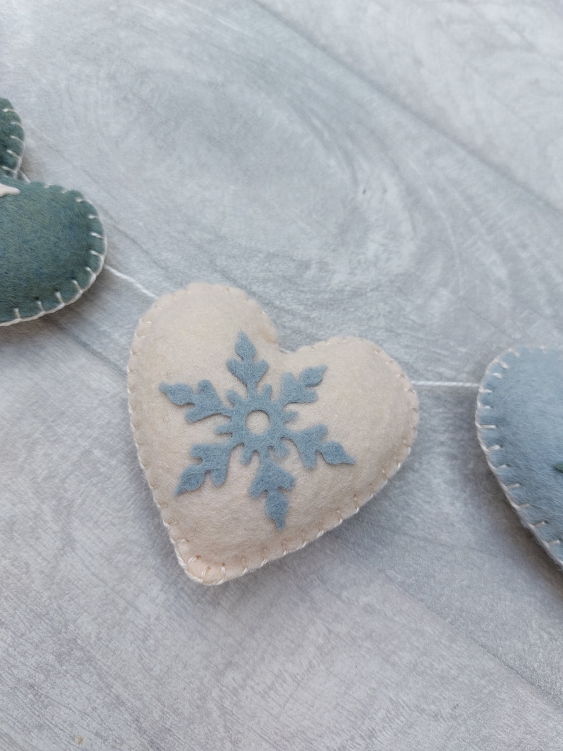 May include: A festive garland featuring felt hearts in cream and teal, stitched with white thread. Each heart is adorned with a light blue snowflake. The garland is strung on a white string, ideal for winter decorations.