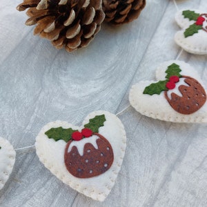May include: A white felt Christmas garland with red and green felt Christmas pudding ornaments. The garland is strung with white thread and has a pine cone in the background.